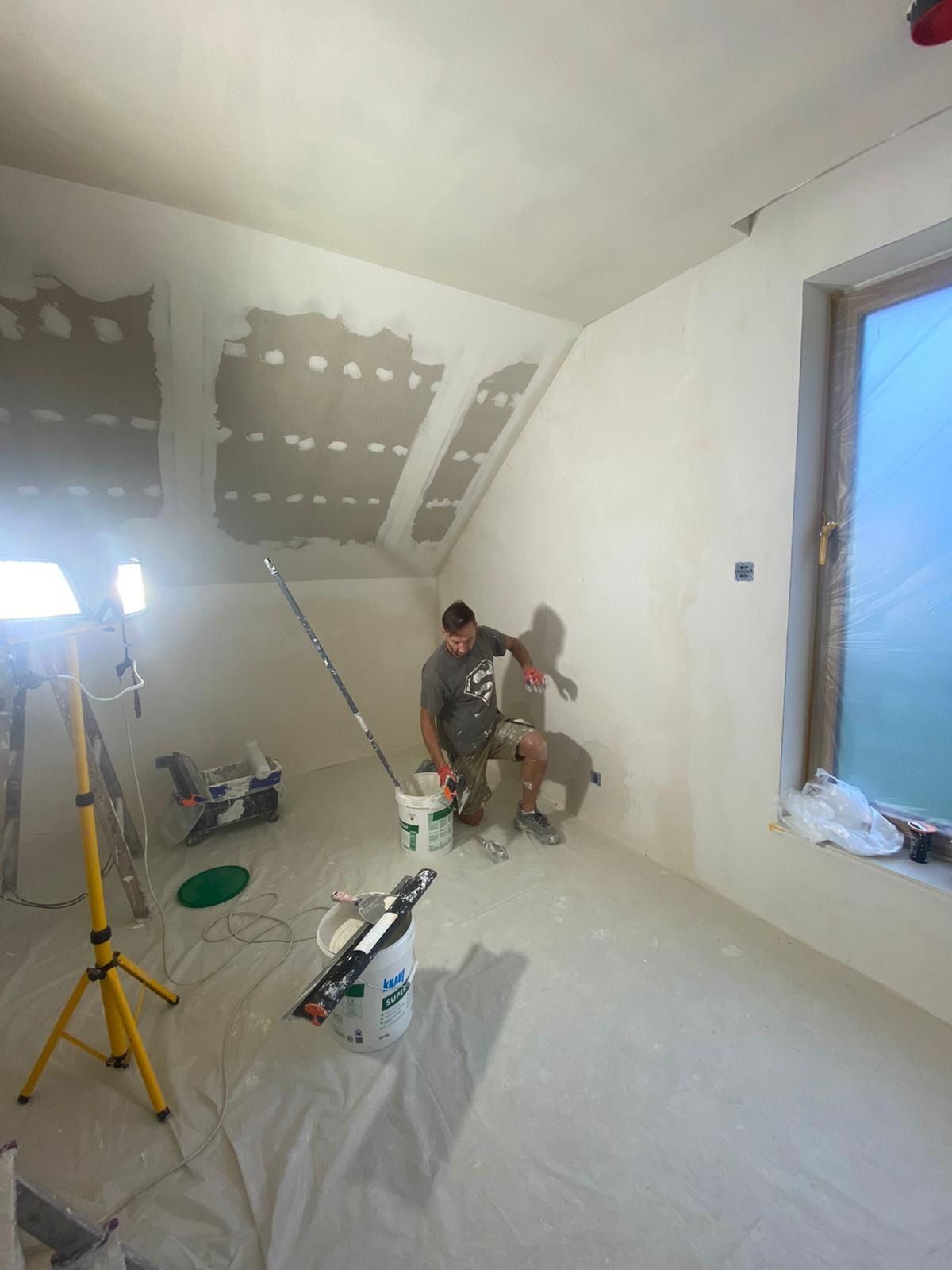 Pracownik w trakcie nakładania gładzi na ścianę poddasza, widoczne narzędzia, wiaderka z masą, lampa oświetleniowa, okno zabezpieczone folią, ściany częściowo pokryte gładzią, sufit z widocznymi...