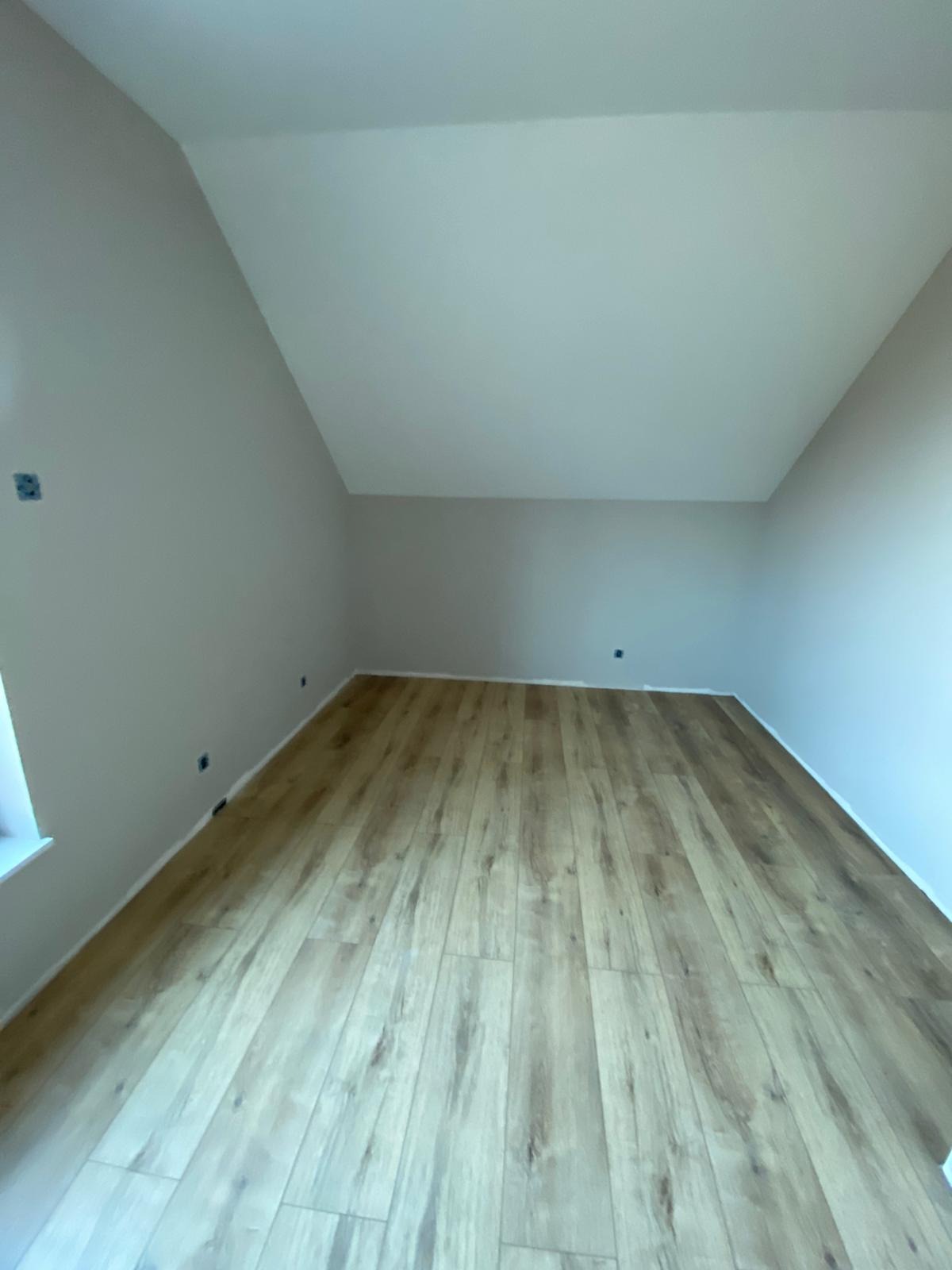 Puste pomieszczenie z nową podłogą laminowaną o wzorze drewna, świeżo pomalowane ściany na jasny odcień szarości, widoczne puszki elektryczne.