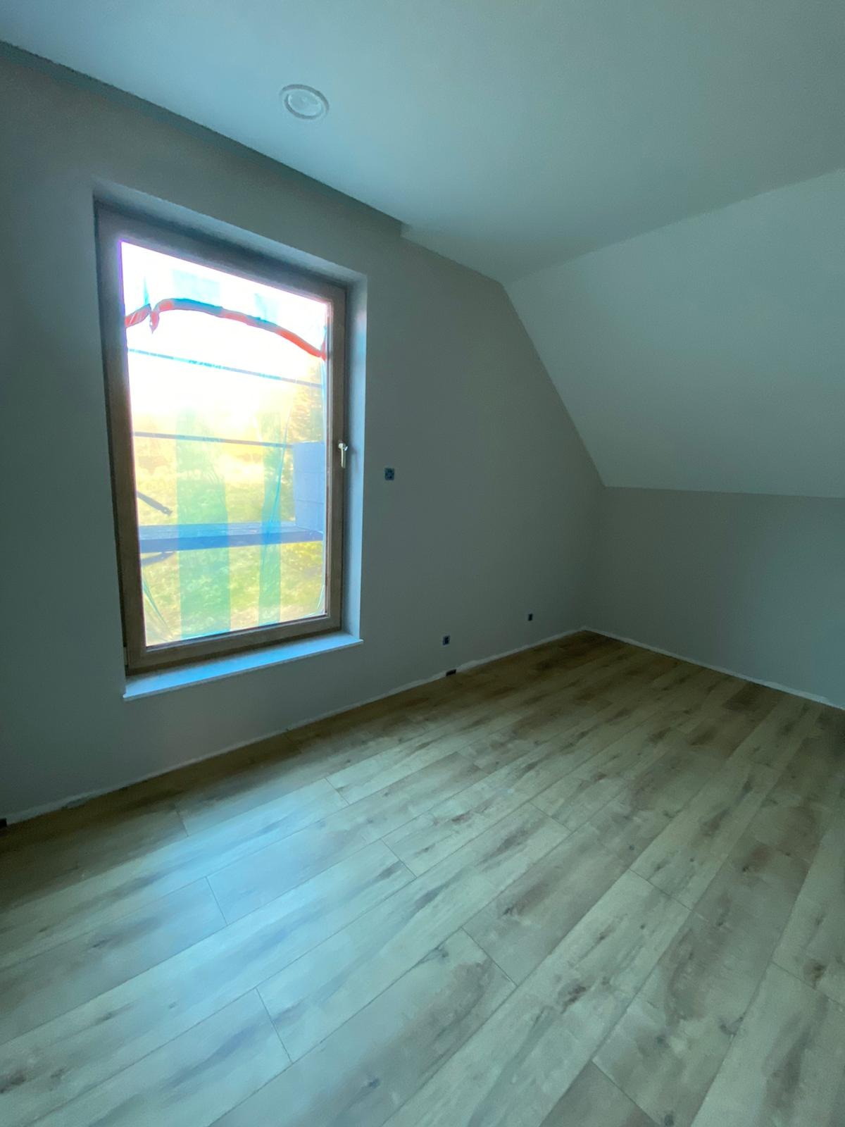 Pokój na poddaszu po remoncie, z drewnianą podłogą, świeżo pomalowanymi szarymi ścianami i oknem zabezpieczonym folią malarską.