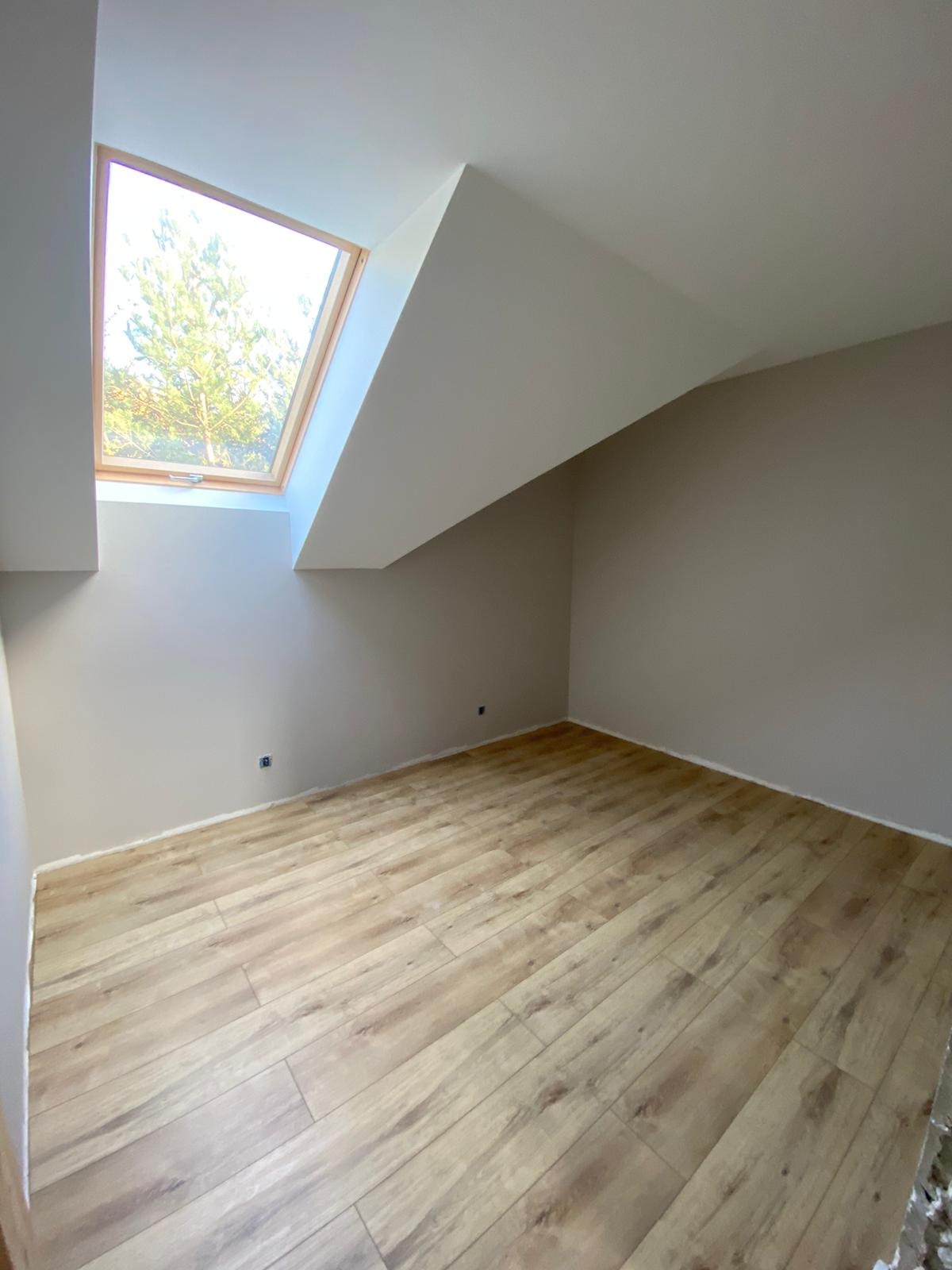 Poddasze z oknem dachowym i panelami podłogowymi w odcieniach beżu, świeżo po malowaniu ścian na jasny kolor, widoczne gniazdko elektryczne.