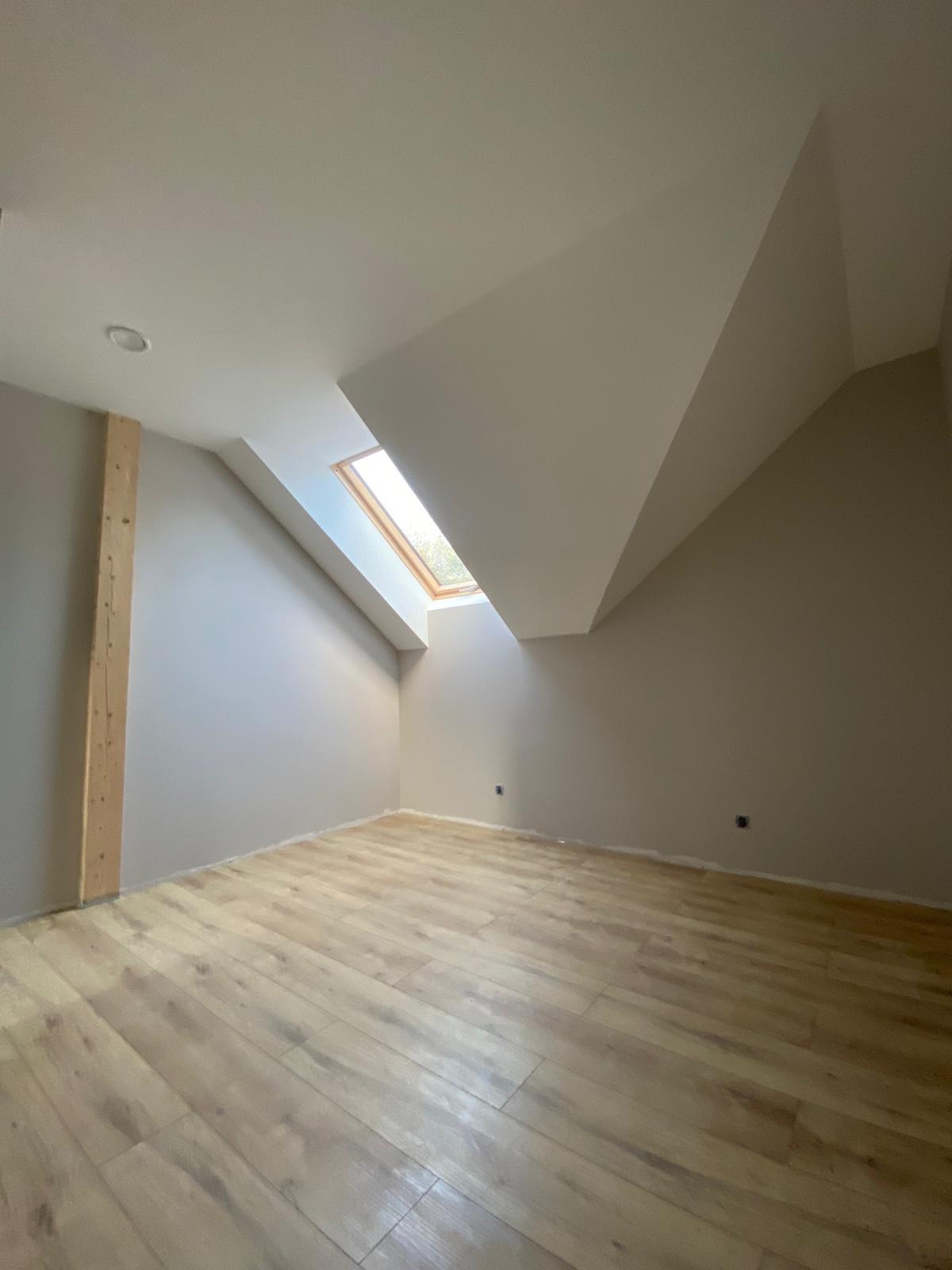 Poddasze po odświeżeniu: pomalowane ściany w odcieniach szarości, drewniana podłoga, okno dachowe wpuszczające naturalne światło, pionowa drewniana belka przy ścianie.