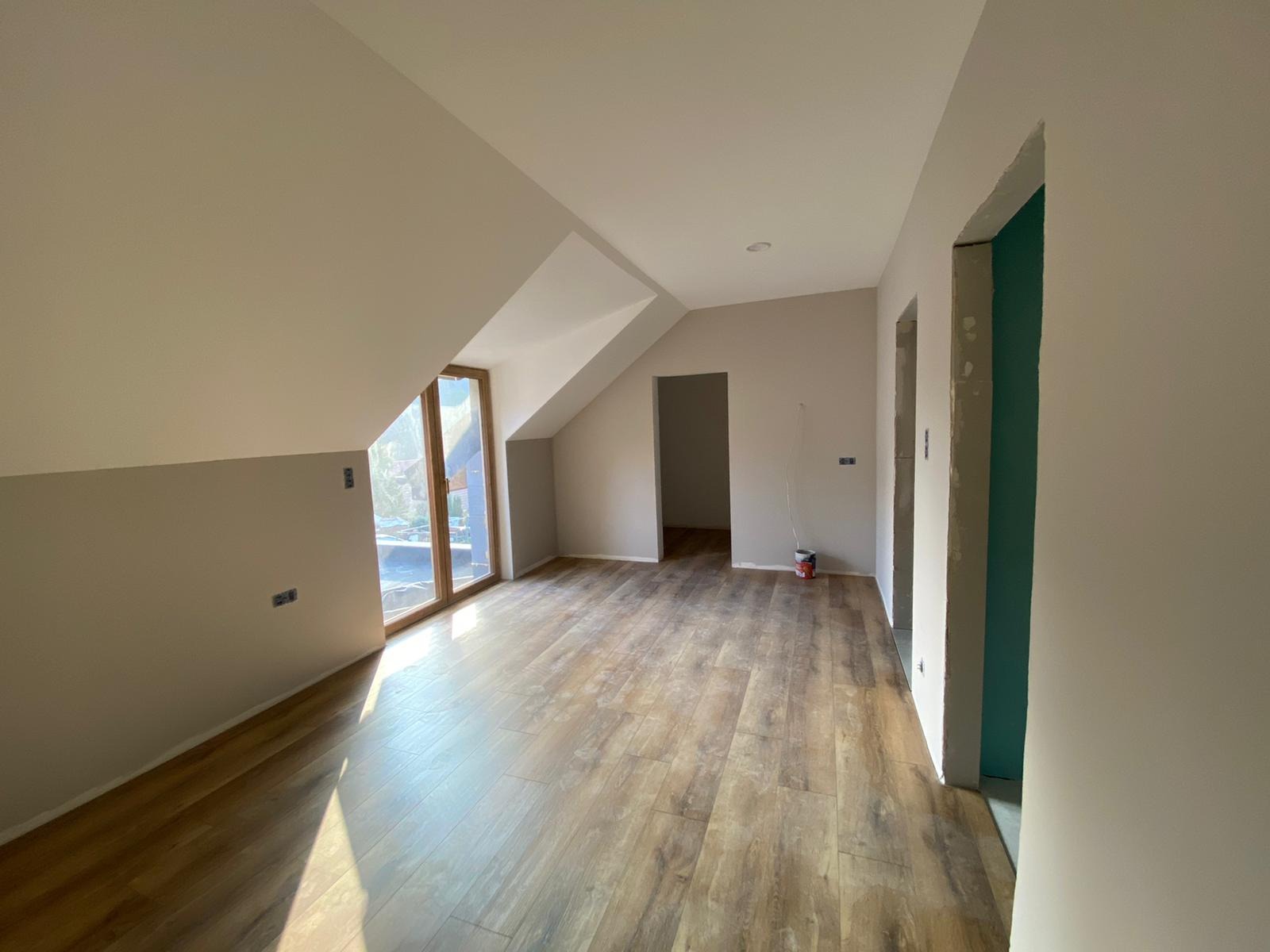 Poddasze z drewnopodobną podłogą, świeżo pomalowanymi ścianami w odcieniach beżu, dużym oknem i widocznymi śladami prac wykończeniowych, w tym niedomalowane fragmenty i wiaderko farby w rogu.