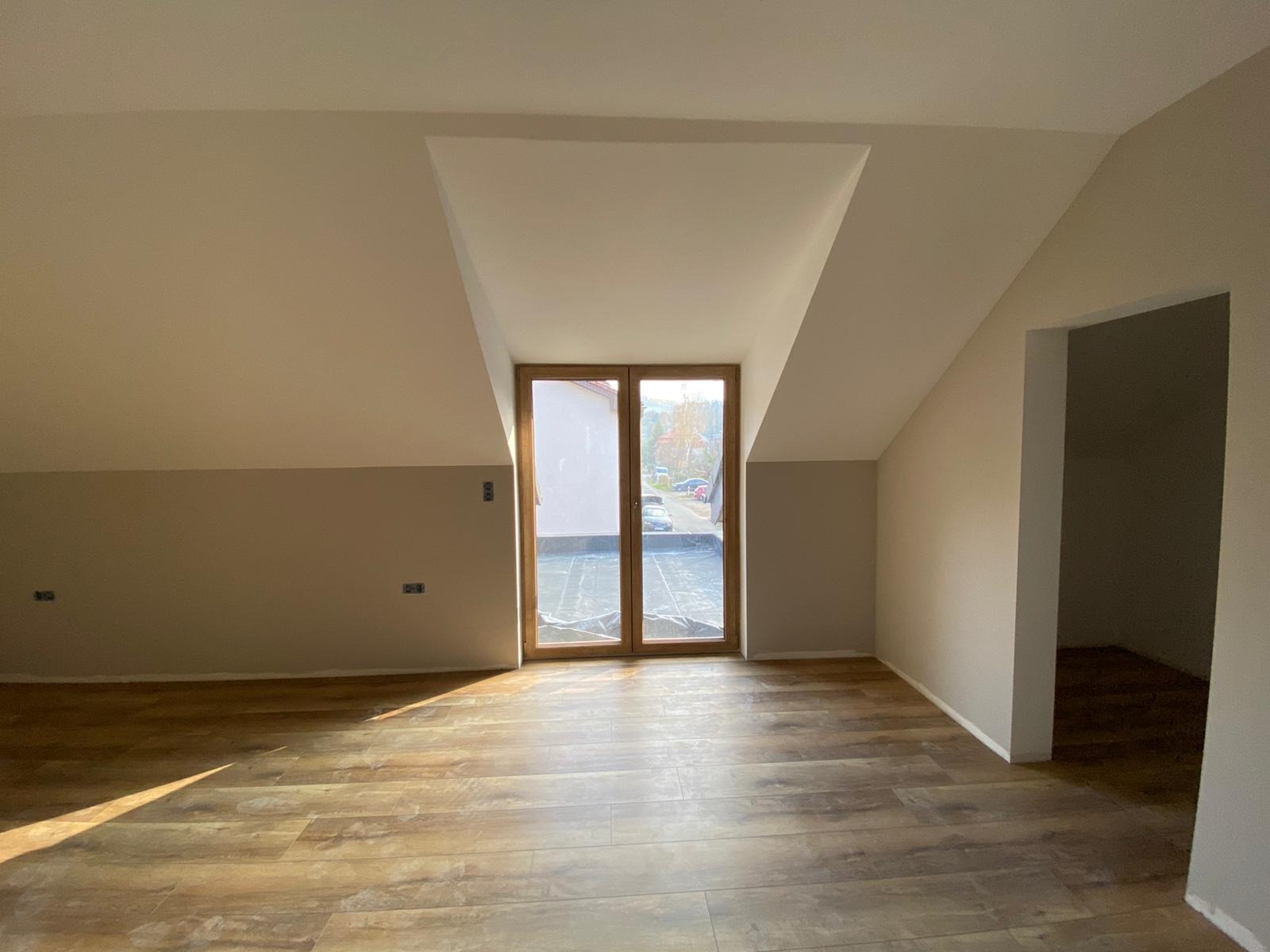 Pokój na poddaszu po odświeżeniu, z drewnopodobną podłogą, dwukolorowymi ścianami (góra kremowa, dół beżowy) i oknem balkonowym z widokiem na zewnątrz.