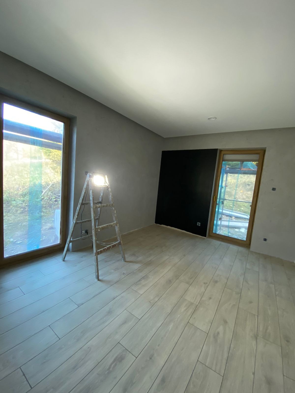 Pokój po świeżo zakończonych pracach, widoczne gładkie ściany w odcieniach szarości, drewniana podłoga, okno z widokiem na zieleń, drabina malarska z lampą halogenową i czarna ściana.