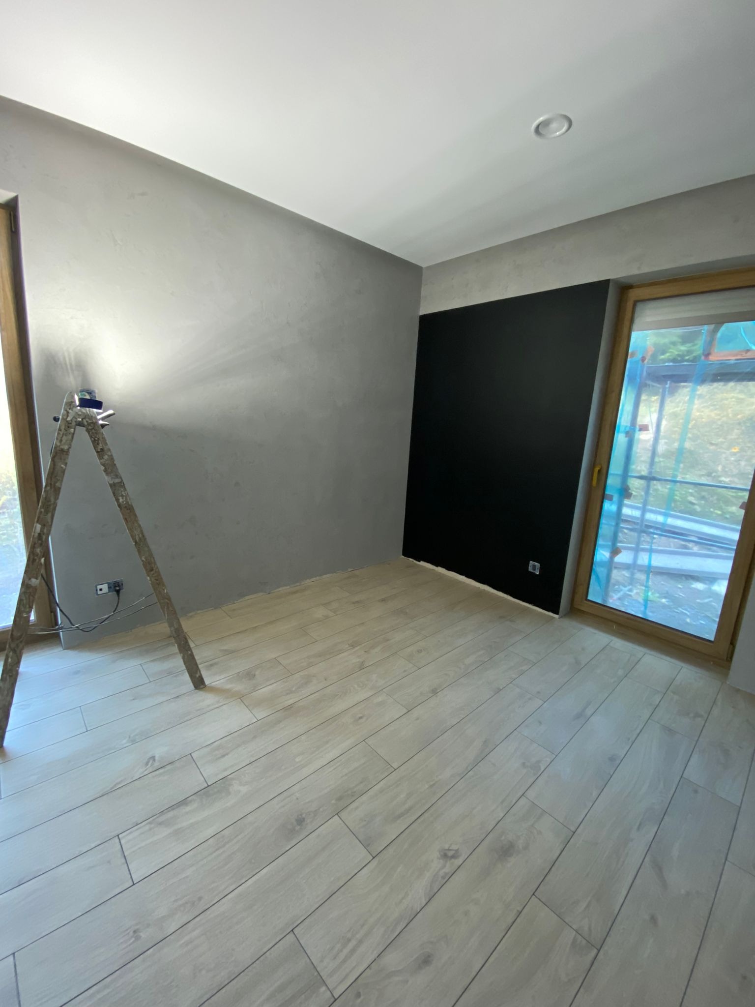 Pokój po częściowym odnowieniu: szara ściana z efektem dekoracyjnym, czarny fragment ściany, okno zabezpieczone folią, drewniana drabina z lampą roboczą, panele podłogowe imitujące drewno.