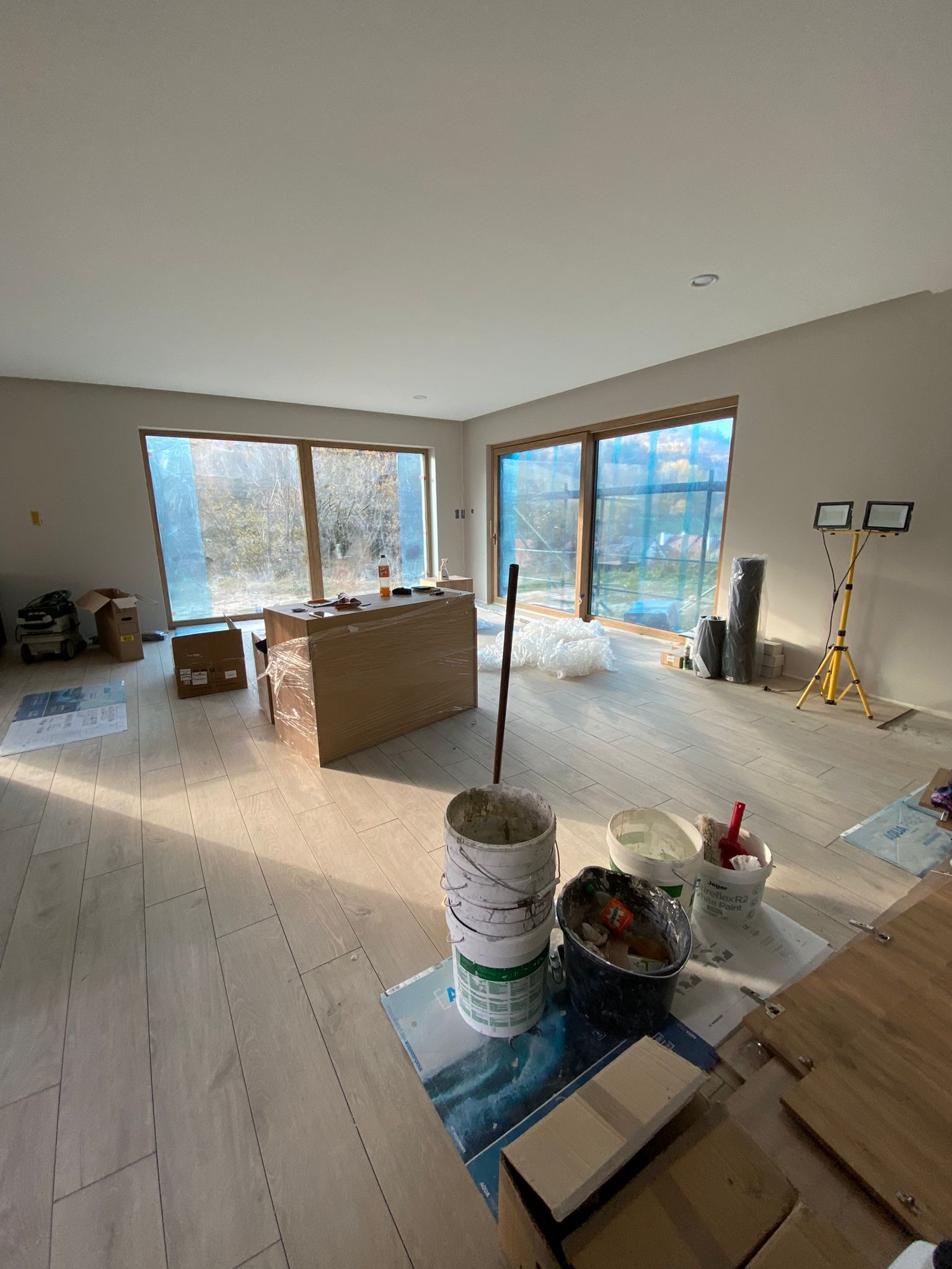 Wnętrze w trakcie remontu z drewnopodobną podłogą, zabezpieczonymi oknami, wiaderkami z farbą i szpachlą oraz profesjonalnym oświetleniem na statywie.