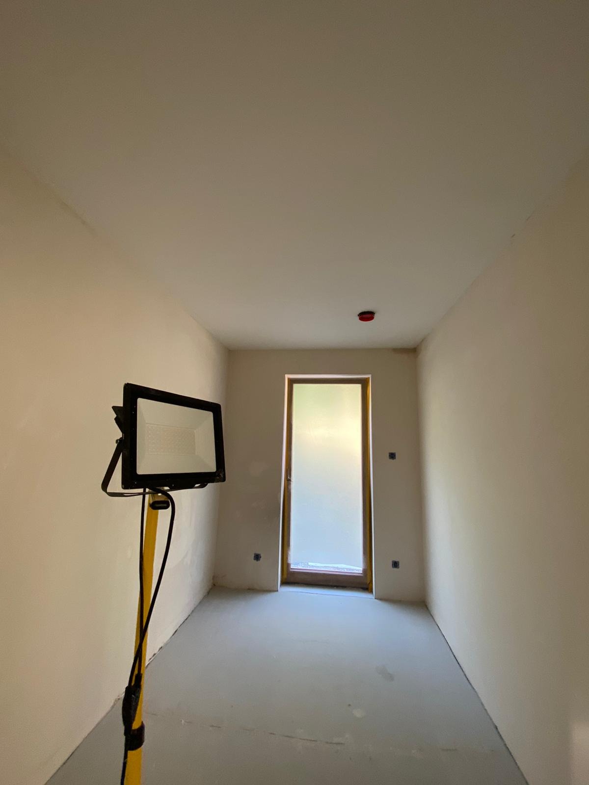 Pokój w trakcie prac przygotowawczych, podłoga zabezpieczona folią, ściany i sufit w jasnym odcieniu, widoczna lampa budowlana na statywie oraz osadzone w ścianie puszki elektryczne.