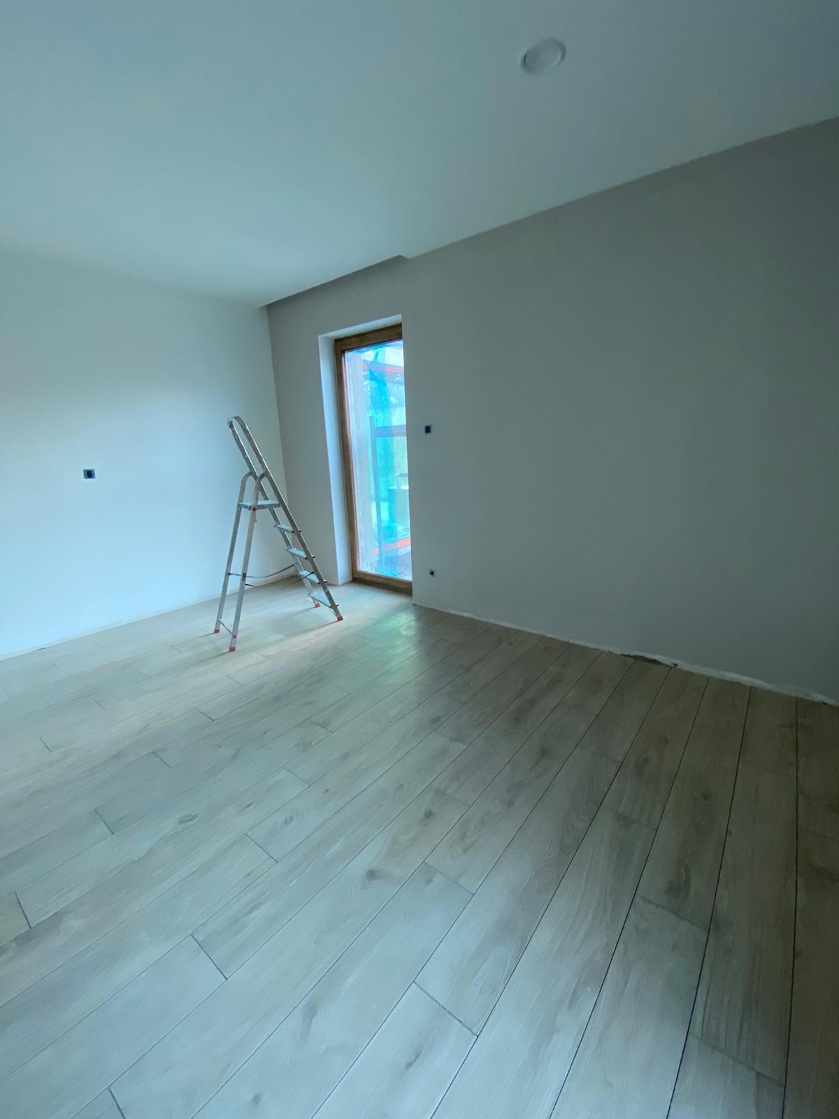 Pokój po świeżym malowaniu, z drewnopodobną podłogą, aluminiową drabiną opartą o ścianę przy oknie zabezpieczonym folią malarską.
