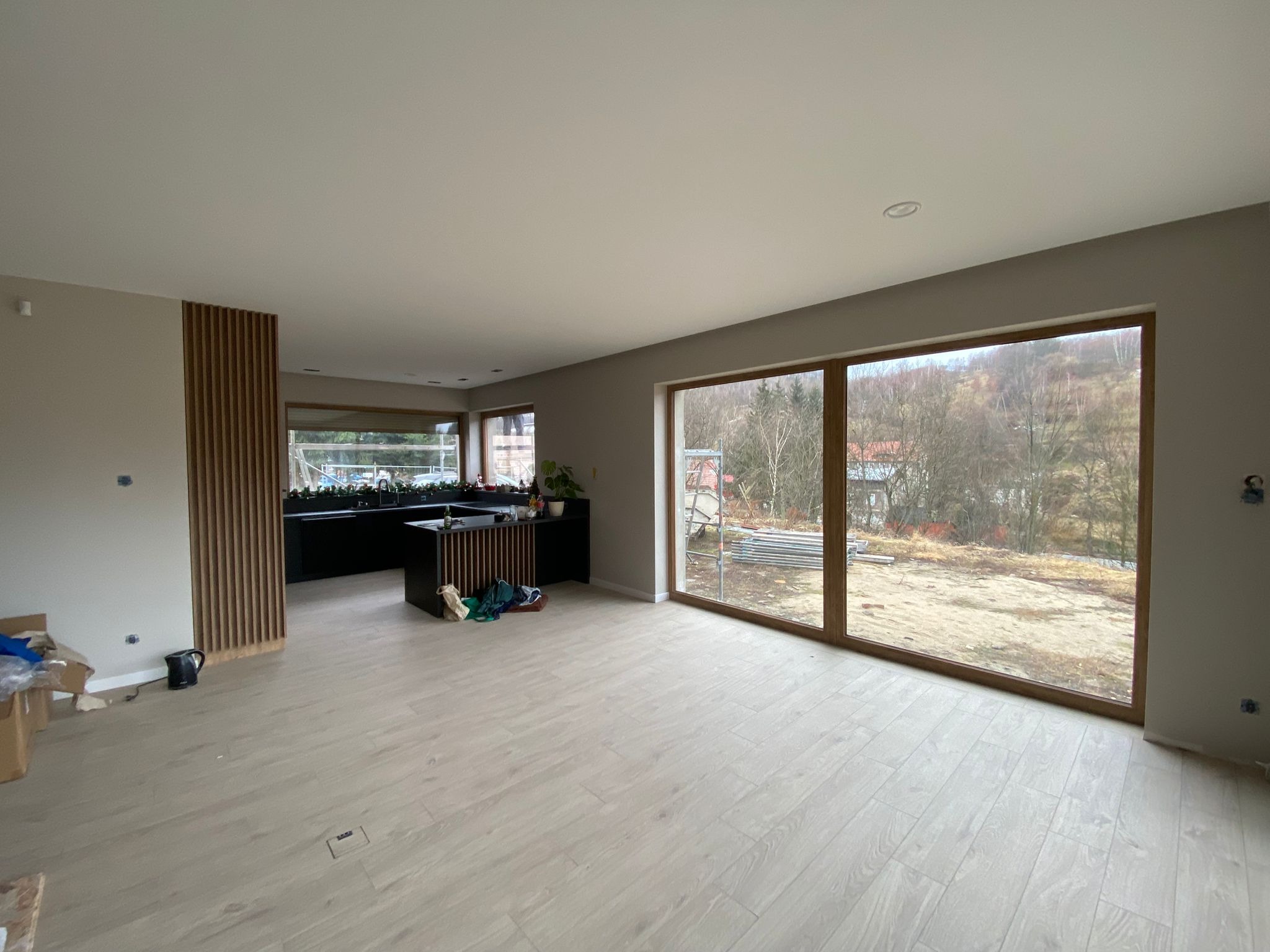 Przestronne wnętrze w stanie deweloperskim z dużym oknem panoramicznym na ogród, widoczna drewniana podłoga, ściany w odcieniach szarości i czarna zabudowa kuchenna z dekoracyjną ścianką...