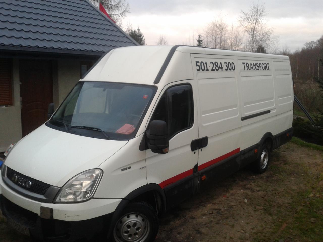 Biały bus Iveco z numerem telefonu i napisem TRANSPORT na boku, zaparkowany na trawie przed domem z ciemnoszarą dachówką.
