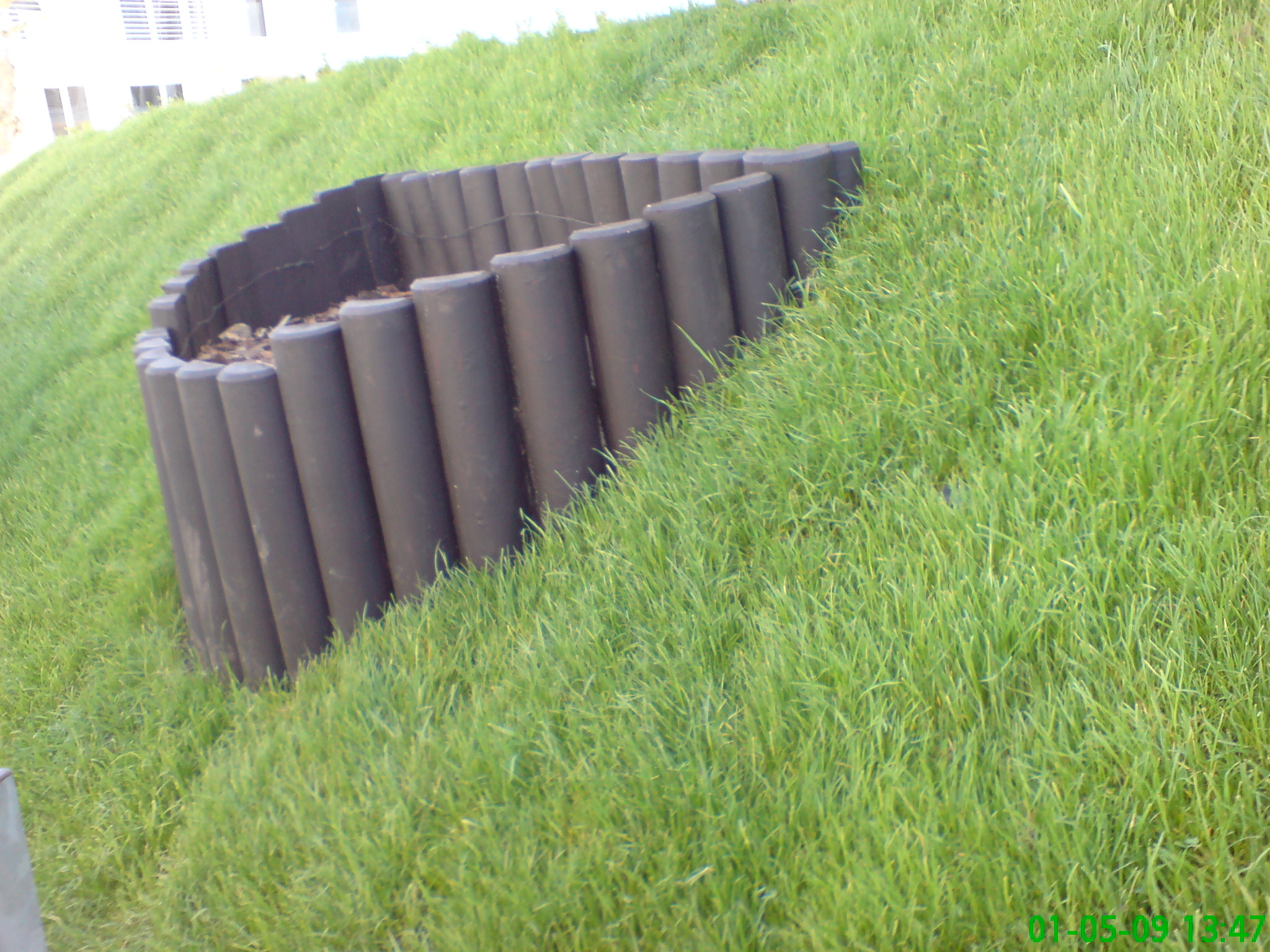 Okrągła rabata obłożona ciemnymi, drewnianymi palisadami na zboczu pokrytym świeżą, zieloną trawą. Widoczny fragment budynku w tle.