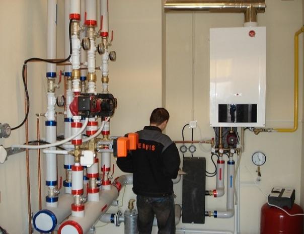 Instalator w trakcie montażu lub serwisu kotła gazowego, widoczne liczne rury, zawory, manometry i osprzęt instalacyjny w pomieszczeniu technicznym.