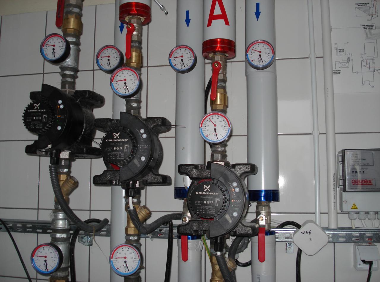 Cztery czarne pompy obiegowe Grundfos podłączone do instalacji z rurami, zaworami z czerwonymi uchwytami i manometrami, na tle białych, kwadratowych płytek ceramicznych.