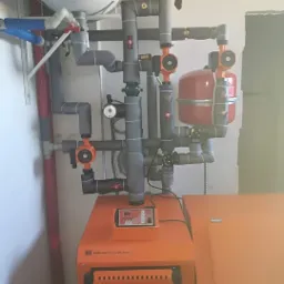 Pomarańczowy piec grzewczy Termo-Tech z podłączonymi szarymi rurami, zaworami i manometrem w surowym wnętrzu.