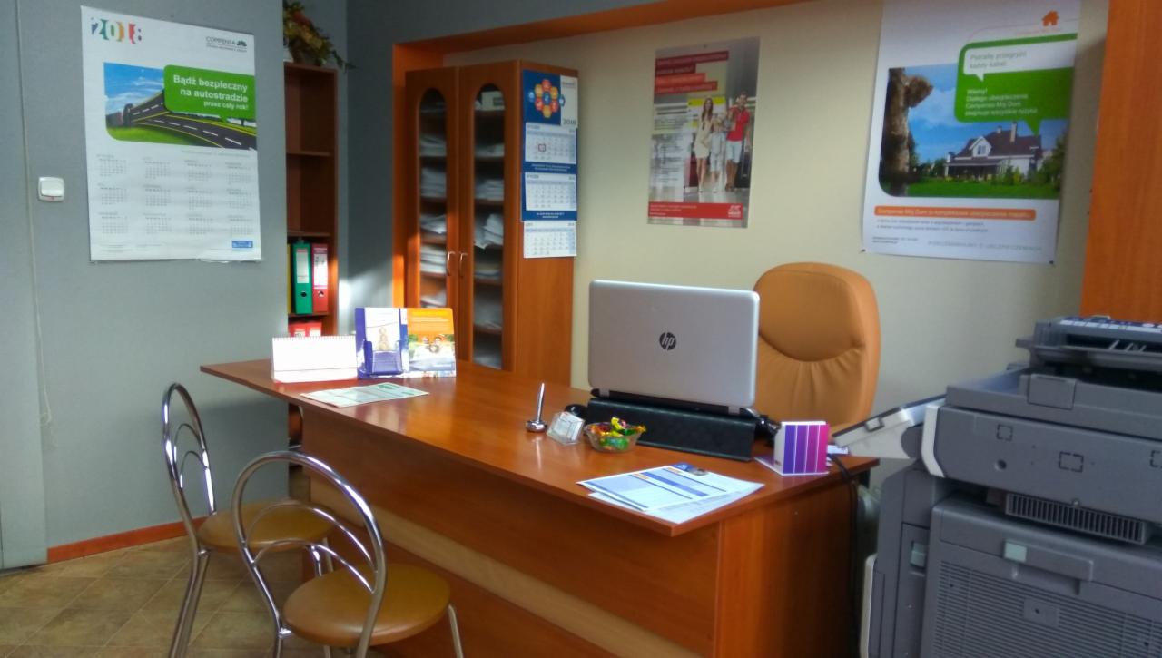 Biuro z biurkiem, laptopem, krzesłami, drukarką i kalendarzami na ścianie, wnętrze firmy ubezpieczeniowej.