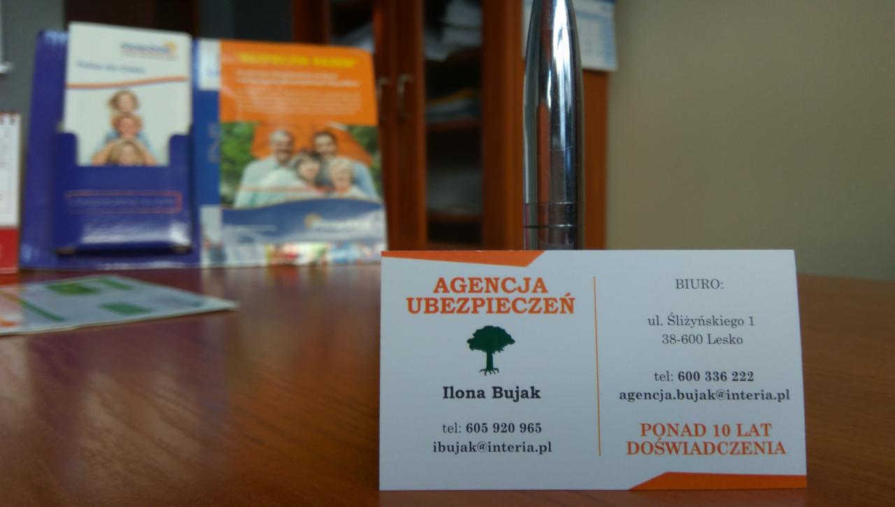 Wizytówka Agencji Ubezpieczeń Ilona Bujak na biurku, z informacjami kontaktowymi i logo drzewa, obok materiałów reklamowych i długopisu.