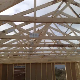 Konstrukcja drewniana dachu domu w trakcie budowy, widoczne wiązary krokwiowe połączone metalowymi płytkami kolczastymi, ściany szkieletowe z płyt OSB, widok na pochmurne niebo.