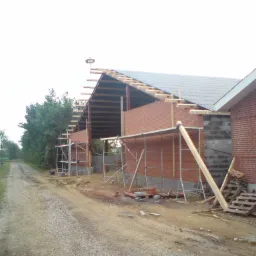 Budowa budynku z czerwonej cegły klinkierowej, widoczne rusztowania, elementy konstrukcji dachu z drewnianymi łatami i częściowo położonymi dachówkami oraz bocianie gniazdo na szczycie dachu.