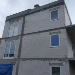 Budynek w trakcie budowy z zamontowanymi oknami z białymi ramami, widoczne bloczki betonowe i niebieska plandeka w dolnej części.