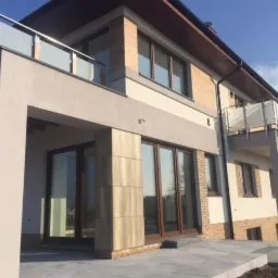 Nowoczesny dom jednorodzinny z balkonami z szklanymi balustradami i dużymi przesuwnymi drzwiami tarasowymi z ciemnego drewna, widok z zewnątrz w słoneczny dzień.