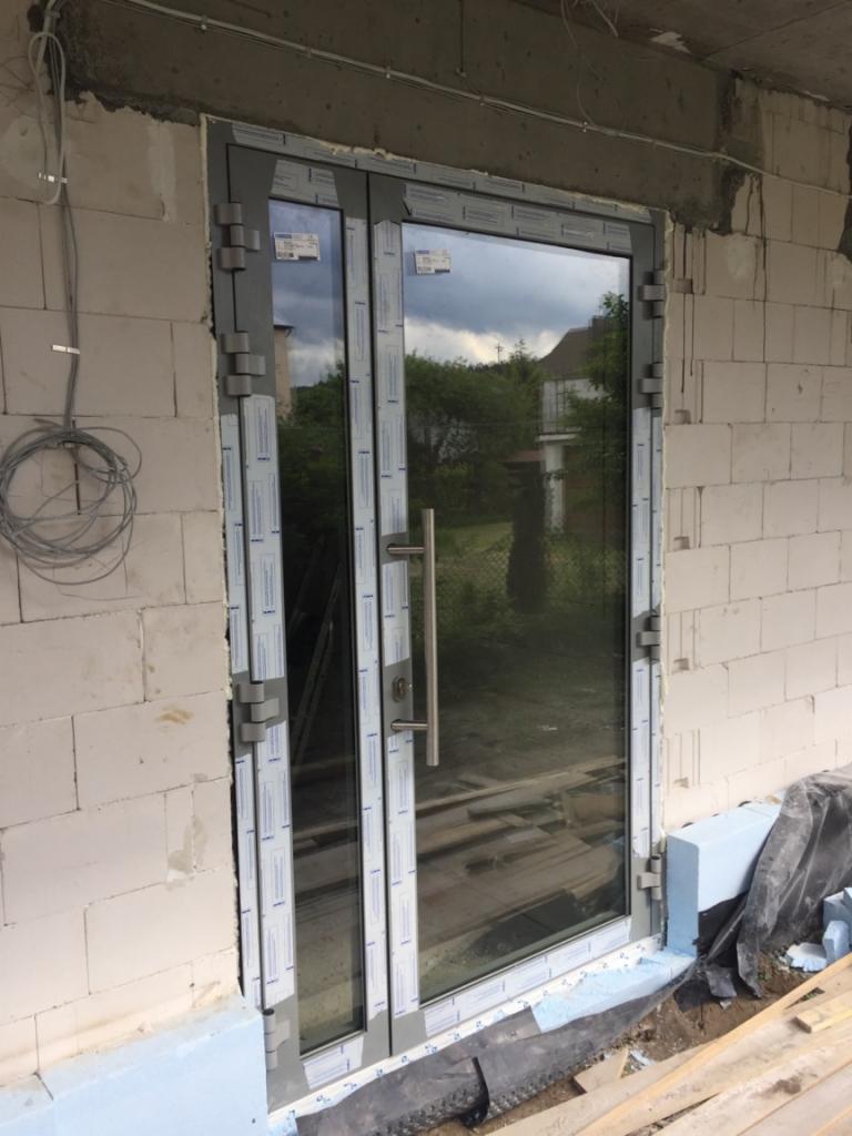 Montaż szklanych drzwi zewnętrznych z szarymi ramami w surowym stanie budynku, widoczne pianka montażowa i przewody elektryczne.