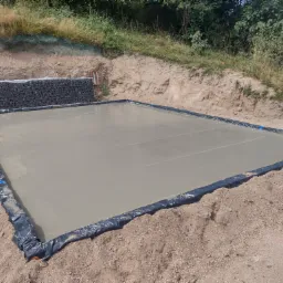 Wylana, świeża płyta betonowa fundamentowa, zabezpieczona czarną folią, widoczna na tle ziemi i muru oporowego z gabionów.
