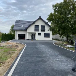 Nowoczesny dom jednorodzinny z ciemnoszarą kostką brukową na podjeździe i chodniku, białe obrzeża, widoczne narzędzia i materiały budowlane, częściowo ukończona realizacja.