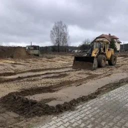 Żółta ładowarka na placu budowy, widoczne ślady opon na piasku, w tle budynek mieszkalny i zarośla, pochmurne niebo.