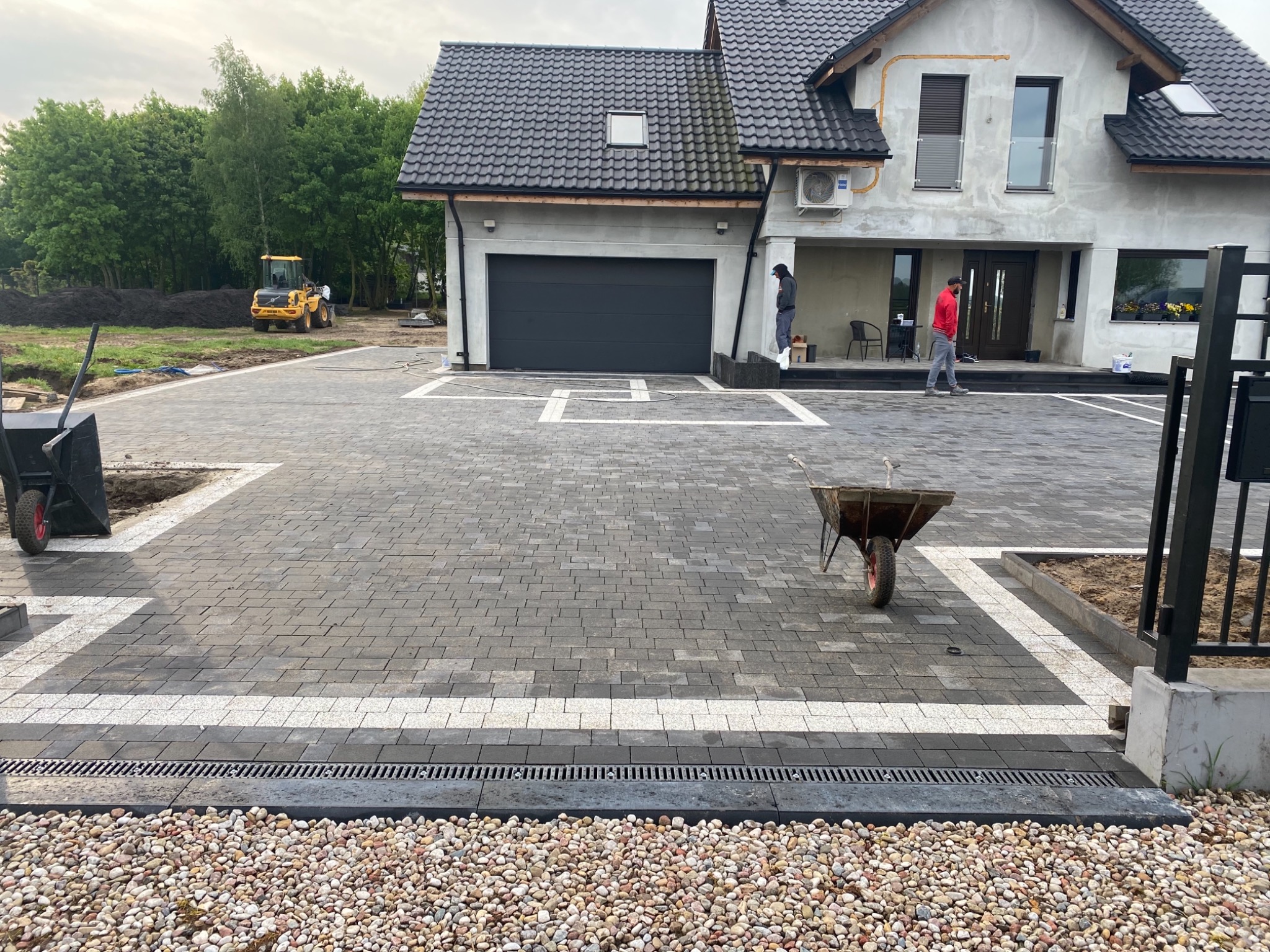 Nowo ułożona, szara kostka brukowa z białymi akcentami na podjeździe przed domem jednorodzinnym, widoczne korytko odwadniające i ozdobny pas z kamyków, w tle koparka i pracownicy.