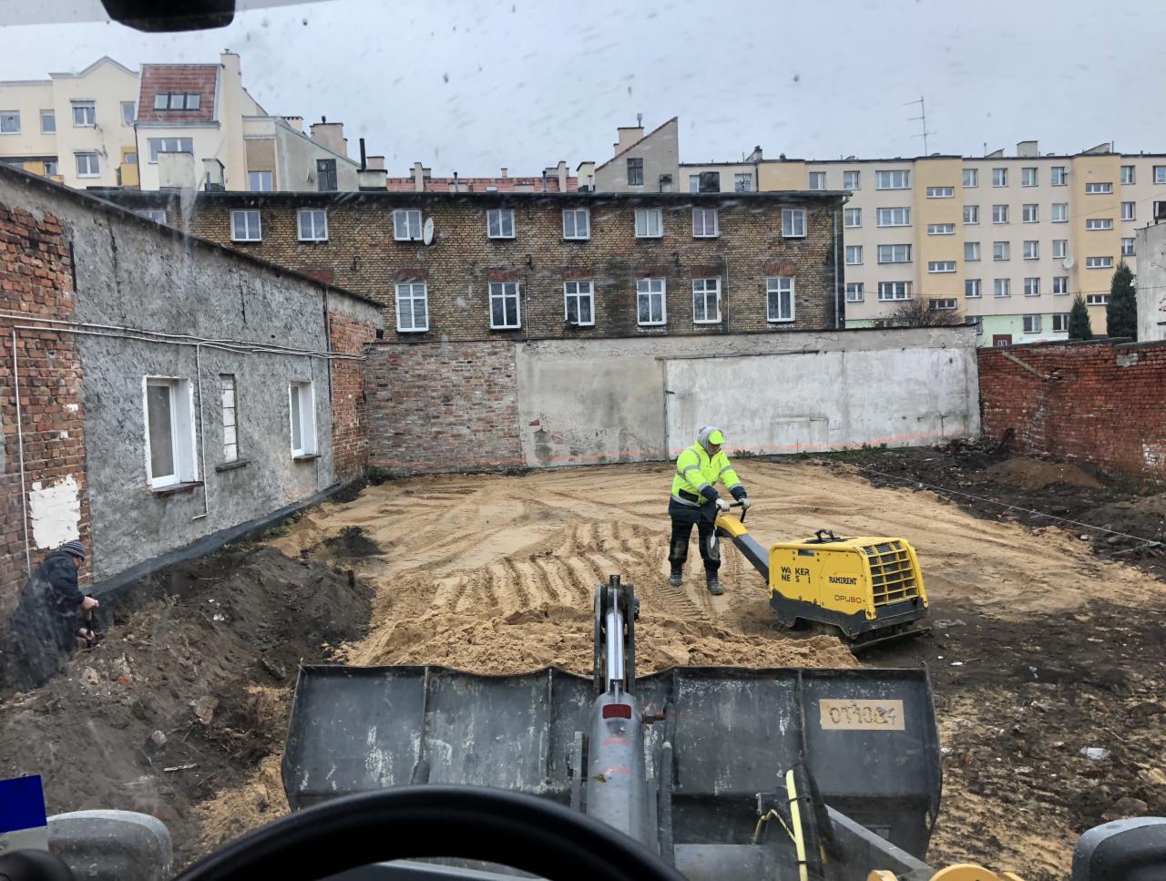 Pracownik w jaskrawej kamizelce używa zagęszczarki na piaszczystym terenie budowy, widok z kabiny ładowarki, w tle zabudowa miejska.