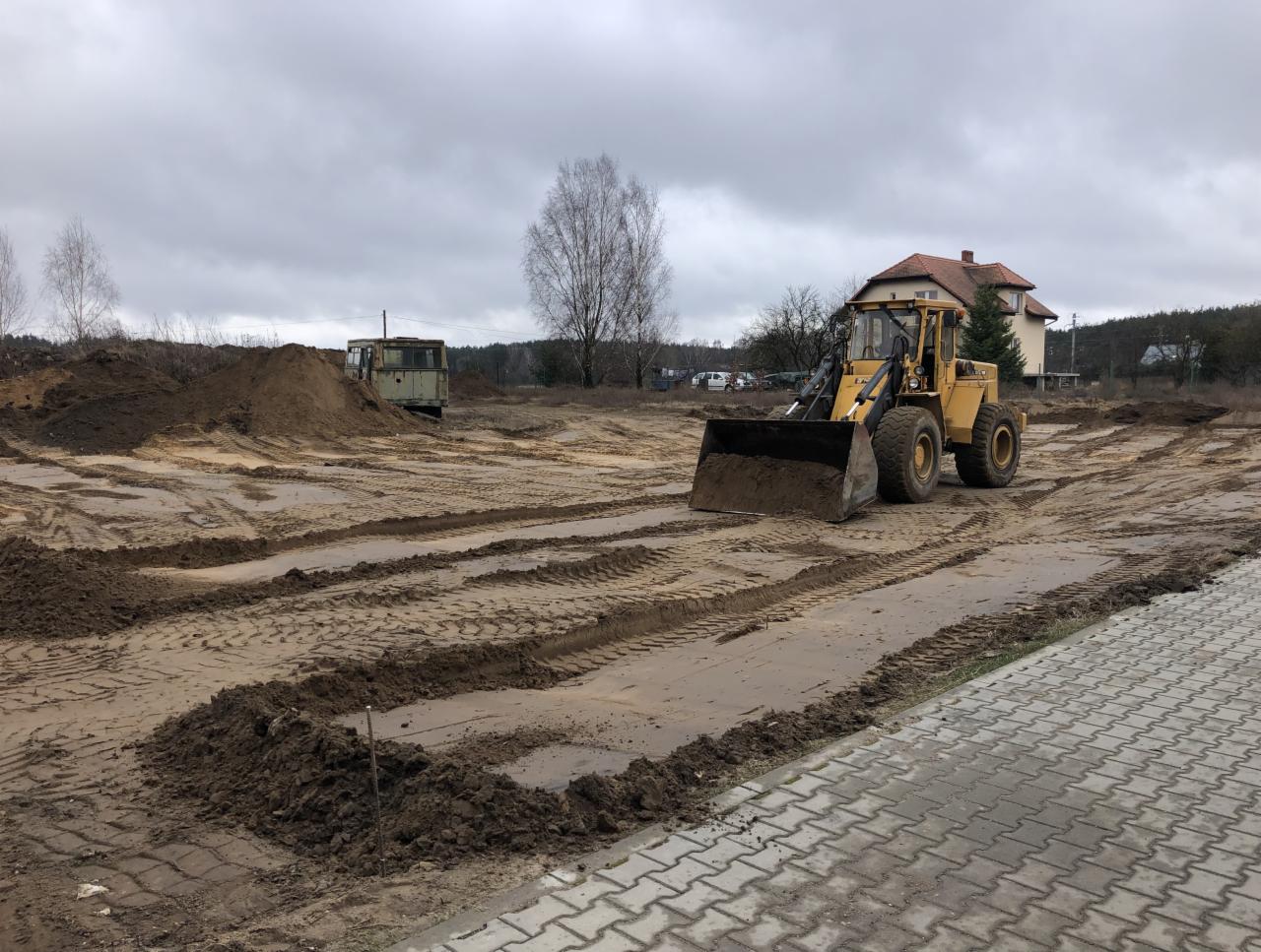 Żółta ładowarka na placu budowy, widoczne ślady opon na piasku, w tle budynek mieszkalny i zarośla, pochmurne niebo.