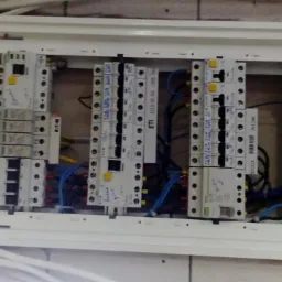 Otwarta skrzynka elektryczna z zamontowanymi bezpiecznikami ETI i Eaton, widoczne niebieskie i czarne przewody oraz brązowy przewód uziemiający, na niektórych bezpiecznikach widoczne odręczne notatki.