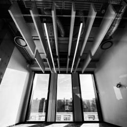 CLS Instalacje Elektryczne Sp. z o.o. - Czarno-białe zdjęcie wnętrza z widokiem na sufit z zamontowanymi panelami akustycznymi i oświetleniem liniowym, widoczne również elementy wentylacji oraz okna z widokiem na budynki.