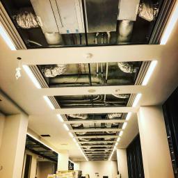 CLS Instalacje Elektryczne Sp. z o.o. - Sufit podwieszany z widocznymi instalacjami wentylacyjnymi i oświetleniem LED w nowym budynku, widok z dołu, w tle materiały budowlane.