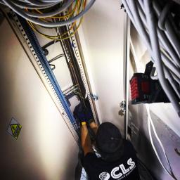 CLS Instalacje Elektryczne Sp. z o.o. - Elektryk w czarnej koszulce z logo firmy CLS Instalacje Elektryczne, w niebieskich rękawicach, instaluje przewody w szafie elektrycznej z widocznymi szynami montażowymi i żółto-zielonymi kablami.