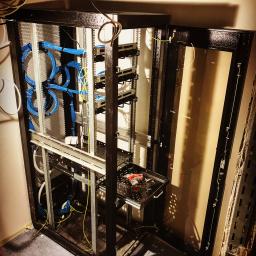 CLS Instalacje Elektryczne Sp. z o.o. - Widok szafy serwerowej z otwartymi drzwiami, wewnątrz uporządkowane kable sieciowe w kolorze niebieskim i żółtym, na wysuwanej półce wewnątrz szafy leży wkrętarka akumulatorowa i klucze