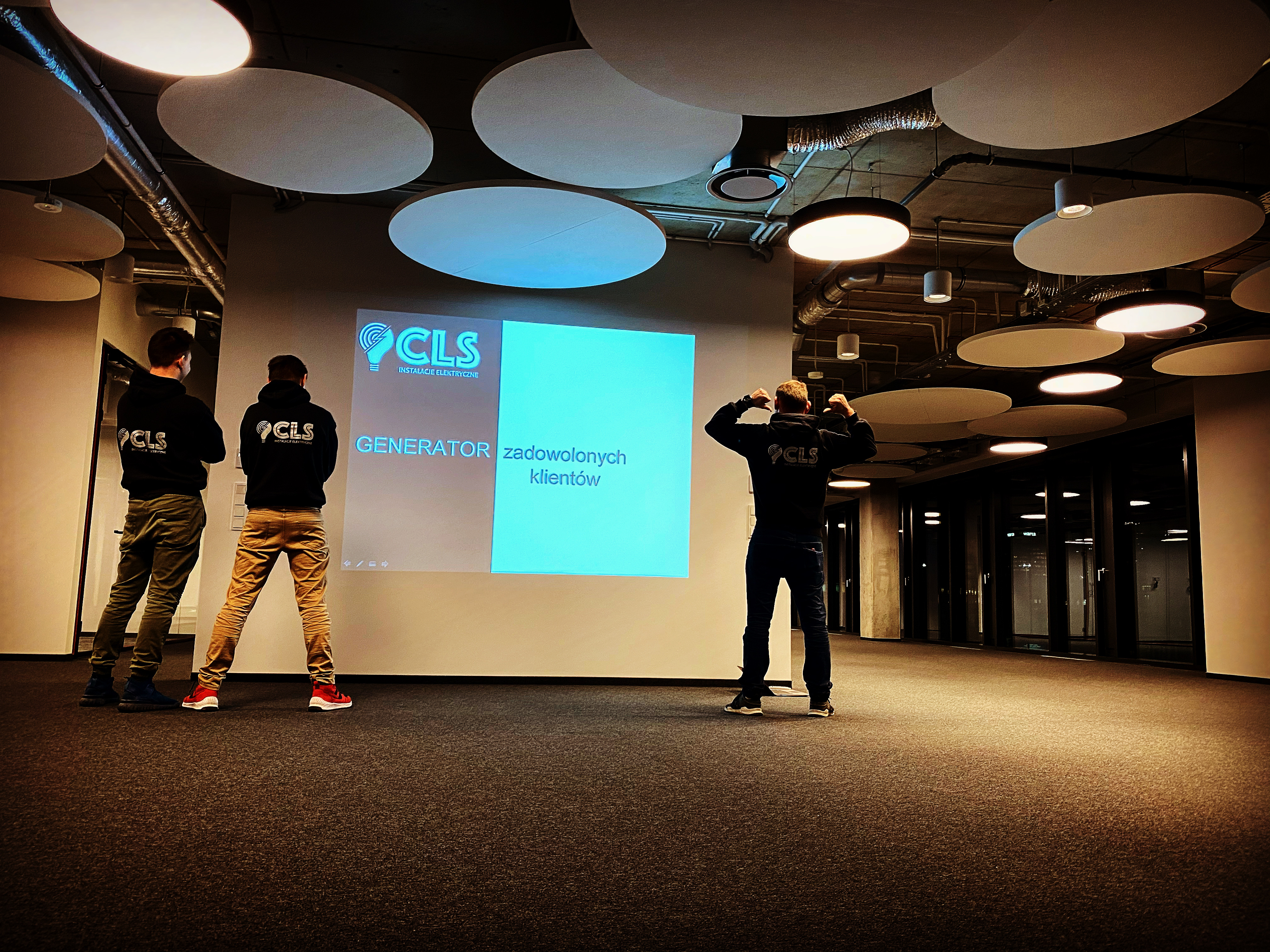 Prezentacja firmy instalacyjnej z Warszawy: trzech mężczyzn w bluzach z logo CLS stoi przed ekranem z napisem 'Generator zadowolonych klientów'.