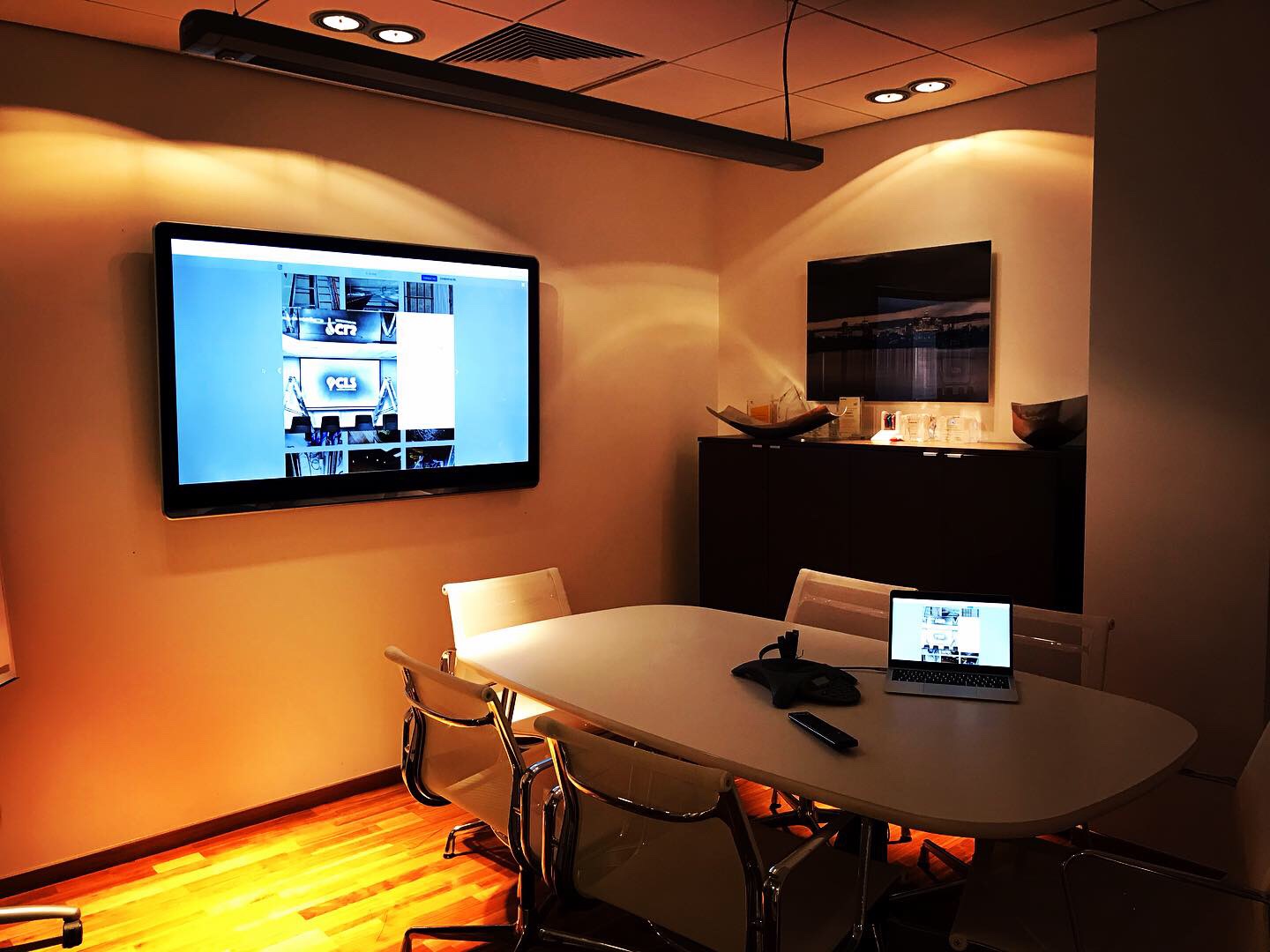 Sala konferencyjna z długim stołem, krzesłami z chromowanymi nogami, laptopem, telefonem konferencyjnym i dużym telewizorem na ścianie wyświetlającym galerię zdjęć.
