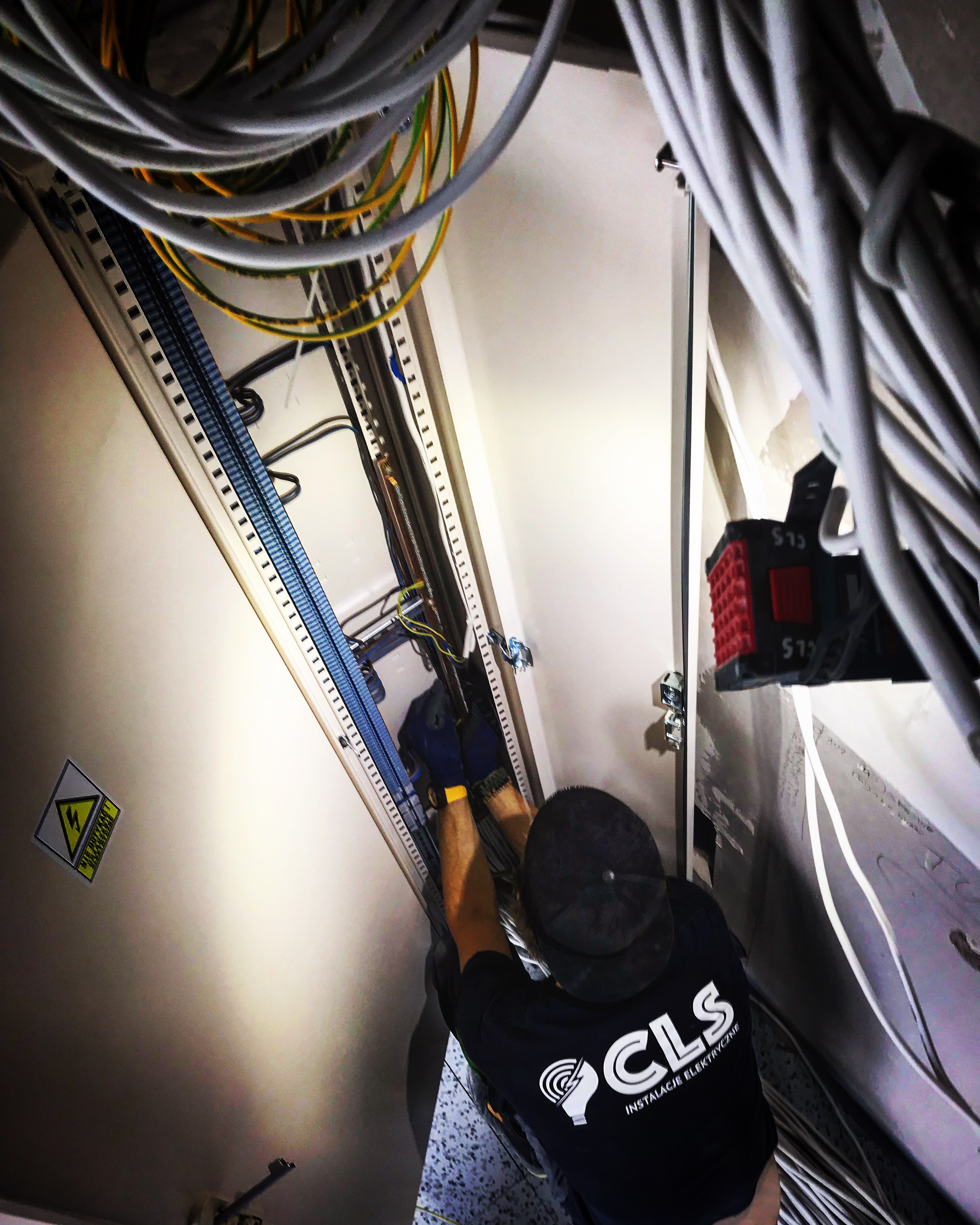 Elektryk w czarnej koszulce z logo firmy CLS Instalacje Elektryczne, w niebieskich rękawicach, instaluje przewody w szafie elektrycznej z widocznymi szynami montażowymi i żółto-zielonymi kablami.