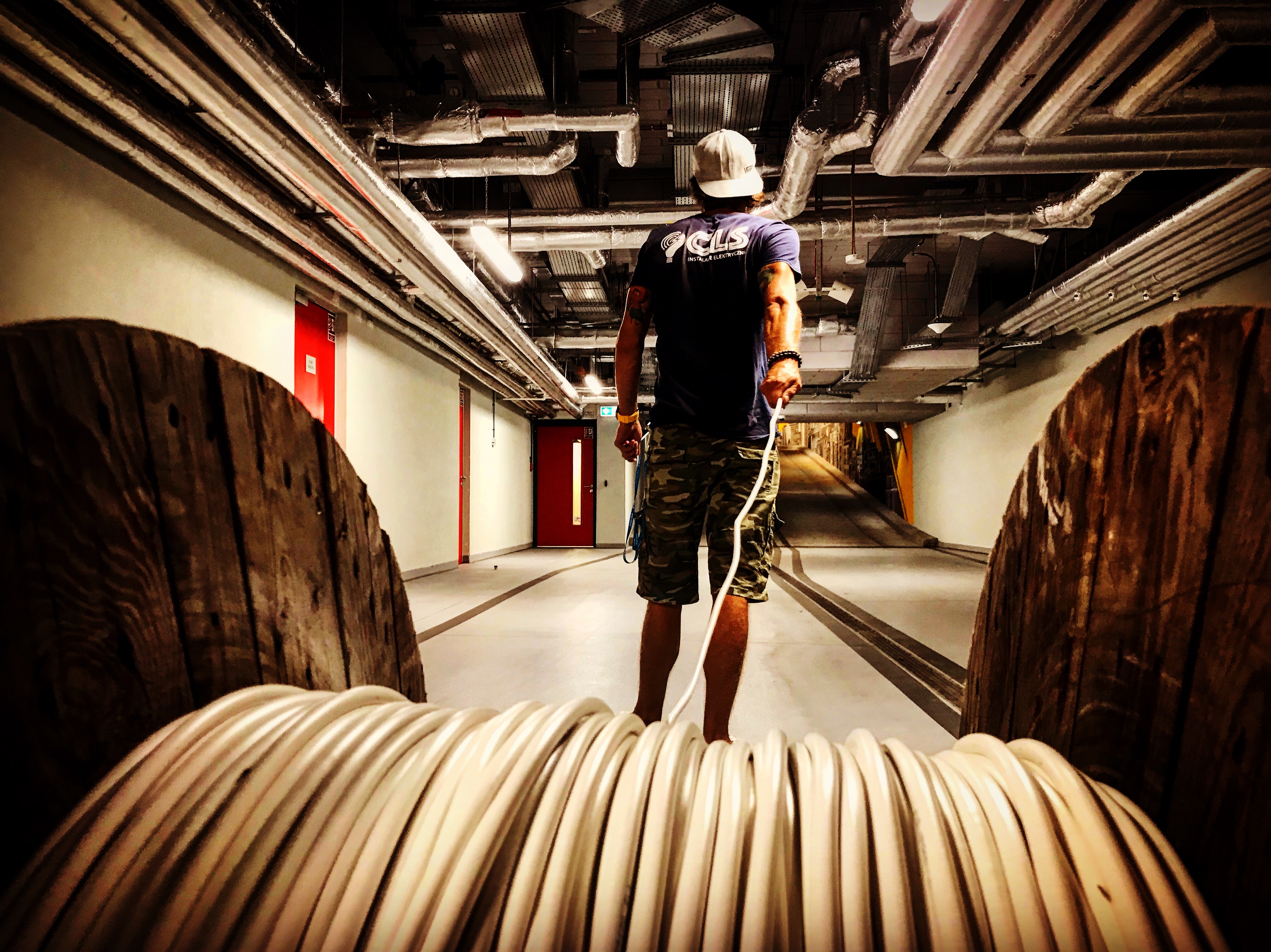Instalator w kasku, z perspektywy bębna z kablem elektrycznym, na tle sufitu z instalacjami w podziemnym garażu.