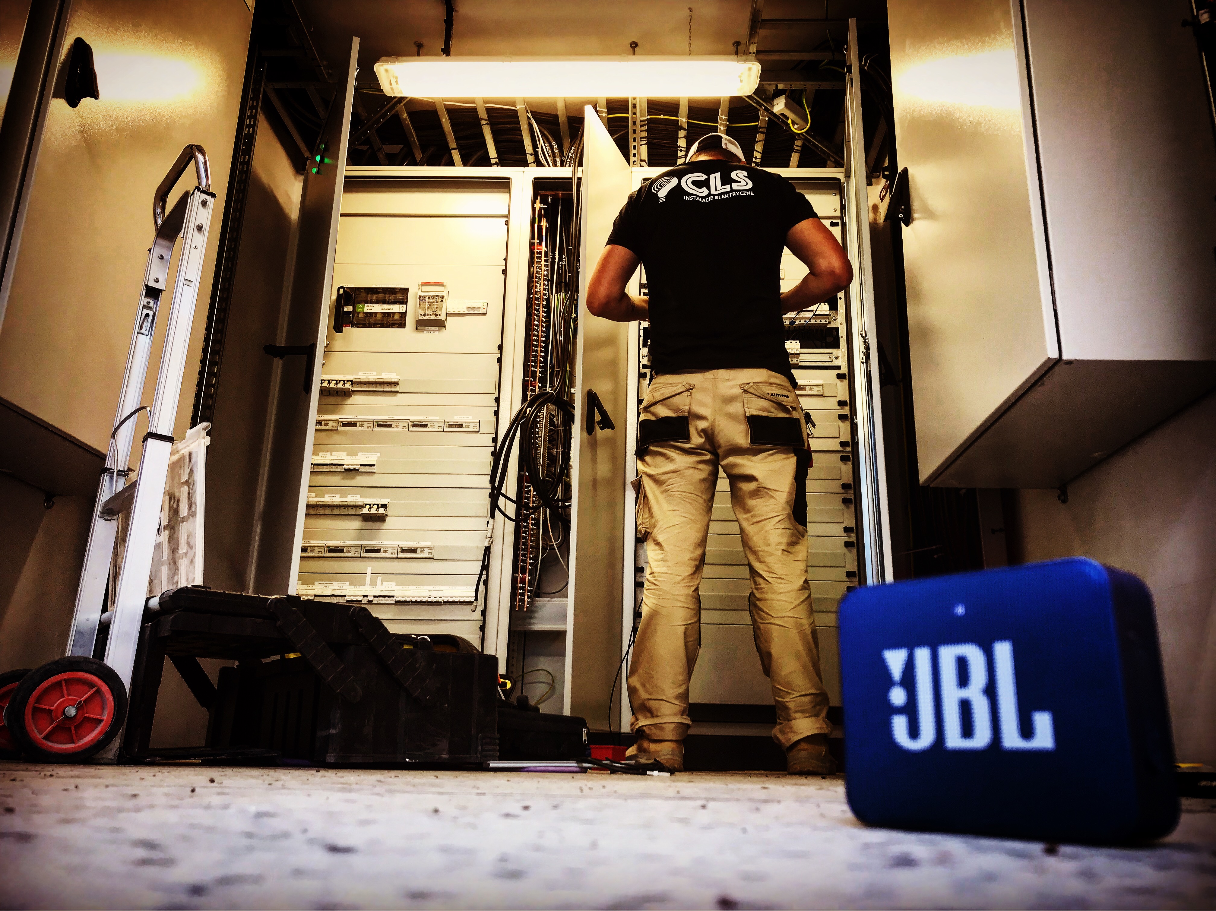 Elektryk w roboczych spodniach, z logo firmy na koszulce, pracuje przy otwartej szafie elektrycznej z rozdzielnicą, obok drabina i głośnik JBL. Nieostre tło.