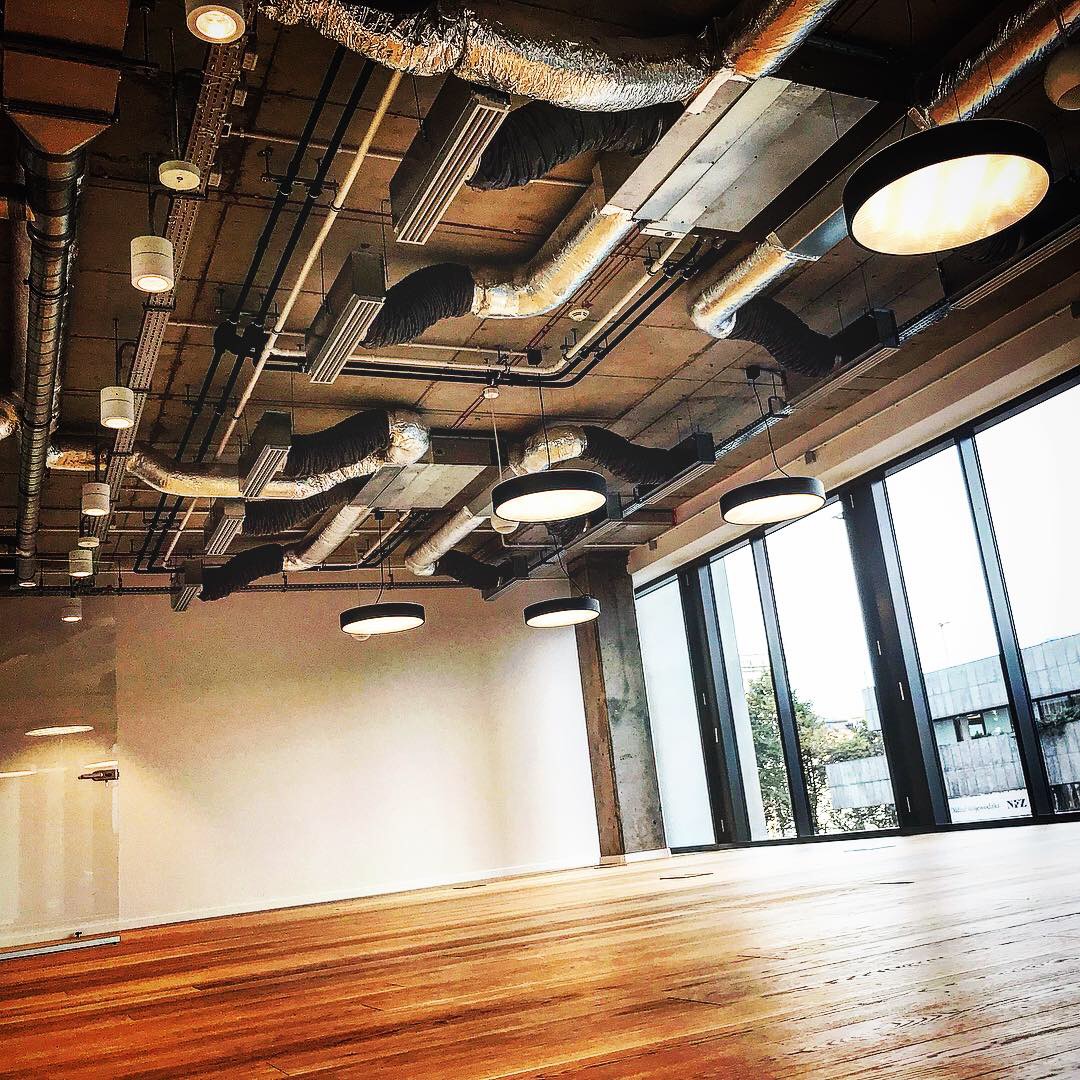 Wnętrze biurowe z widoczną instalacją wentylacyjną pod sufitem, nowoczesnymi lampami wiszącymi i dużymi oknami z widokiem na miasto.