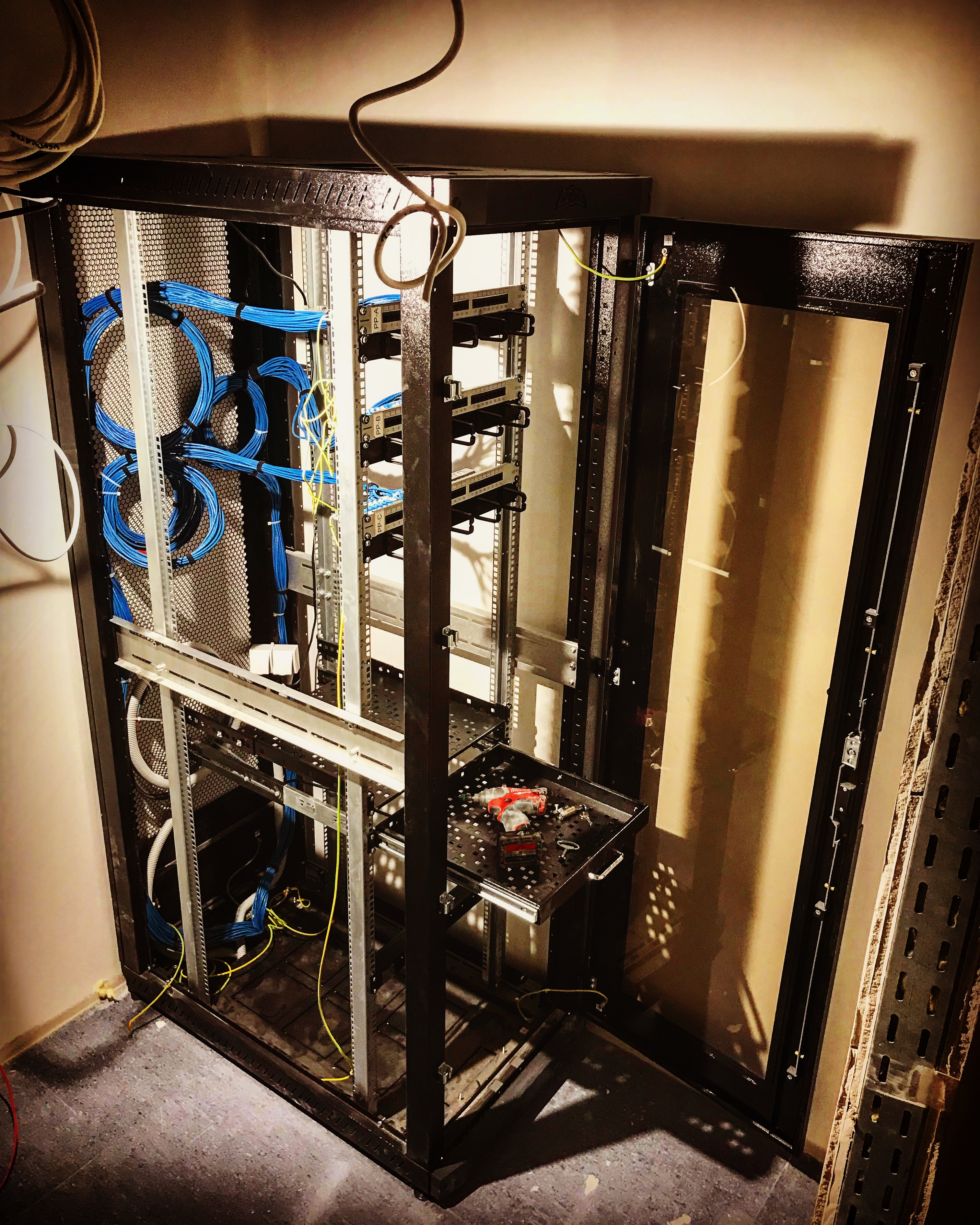 Widok szafy serwerowej z otwartymi drzwiami, wewnątrz uporządkowane kable sieciowe w kolorze niebieskim i żółtym, na wysuwanej półce wewnątrz szafy leży wkrętarka akumulatorowa i klucze