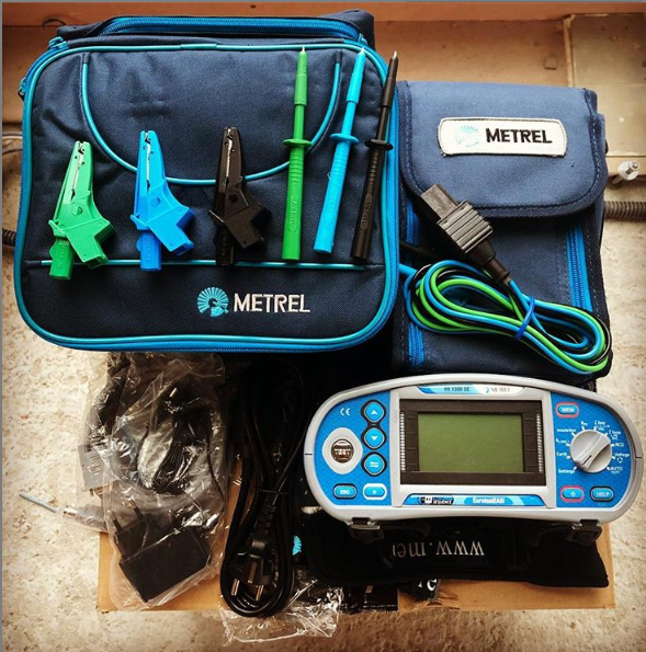 Zestaw narzędzi pomiarowych Metrel: miernik wielofunkcyjny, przewody z krokodylkami i sondami w kolorowych obudowach, zasilacz, instrukcja, umieszczone na kartonowym podłożu.