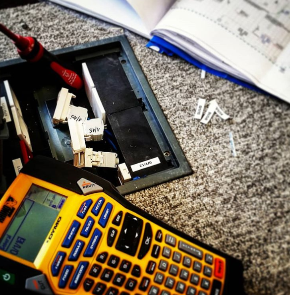 Otwarta puszka podłogowa z kablami i złączkami, oznaczona etykietami, obok leży etykieciarka Dymo, śrubokręt i rozłożony schemat instalacji.