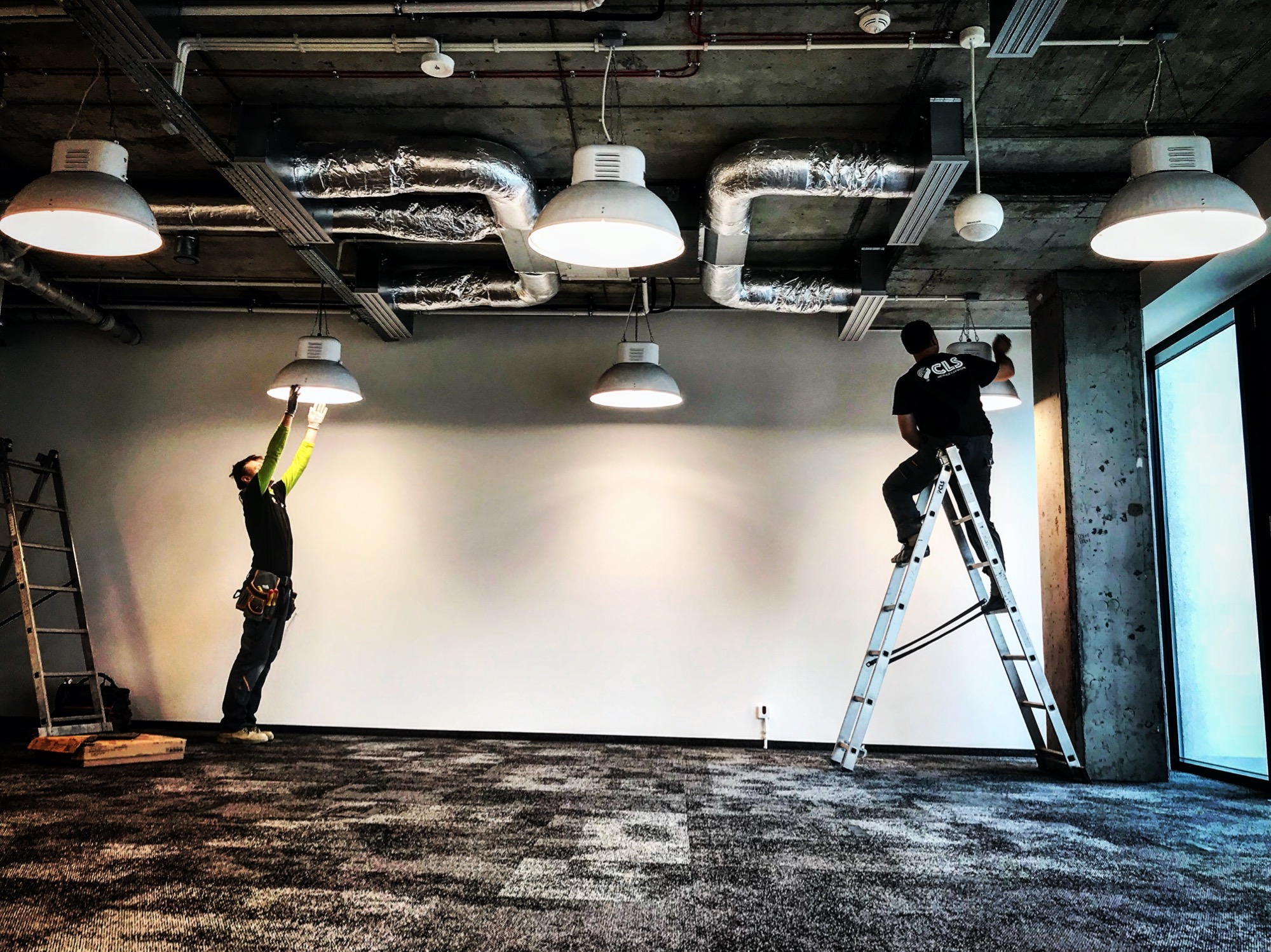 Dwóch mężczyzn na drabinach instaluje przemysłowe lampy wiszące w biurze z widocznymi rurami wentylacyjnymi i betonowym sufitem.