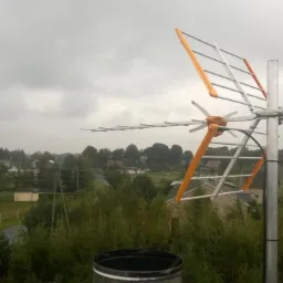 Antena telewizyjna z pomarańczowymi elementami ustawiona na dachu, widok na zalesiony krajobraz pod pochmurnym niebem.