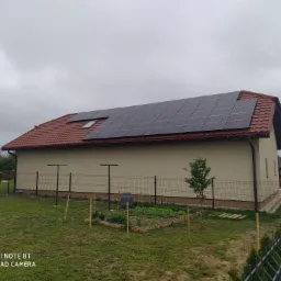 Dom jednorodzinny z panelami fotowoltaicznymi na dachu z czerwonej dachówki, widok z ogrodu z warzywnikiem i młodym drzewkiem, pochmurny dzień.