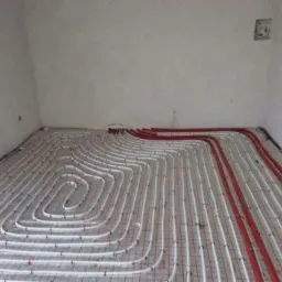 Instalacja ogrzewania podłogowego w trakcie montażu, widoczne białe rurki ułożone spiralnie i czerwone rurki wzdłuż ściany, całość na czarnej folii izolacyjnej w surowym pomieszczeniu.