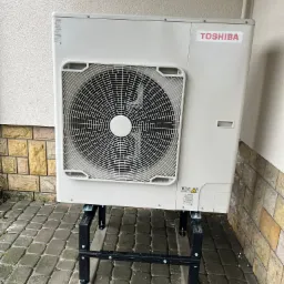 Zewnętrzna jednostka klimatyzacji Toshiba na czarnym, metalowym stelażu, ustawiona na kostce brukowej przy elewacji budynku.